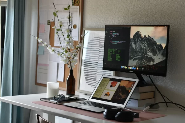 Ein Schreibtisch mit einem Laptop und Monitor, umgeben Accessoires. Das natürliche Licht kommt sanft von der linken Seite.